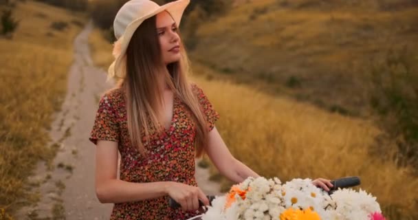 Jeune blonde souriante en chapeau et robe marchant en robe avec vélo et fleurs dans le panier . — Video