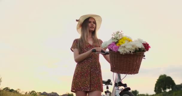 Porter meisje wandelen op de camera in een jurk met bloemen in een mand en een fiets in het veld. — Stockvideo