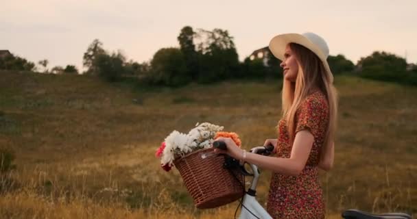 Jonge lachende blonde in muts en jurk wandelen in jurk met fiets en bloemen in mand. — Stockvideo
