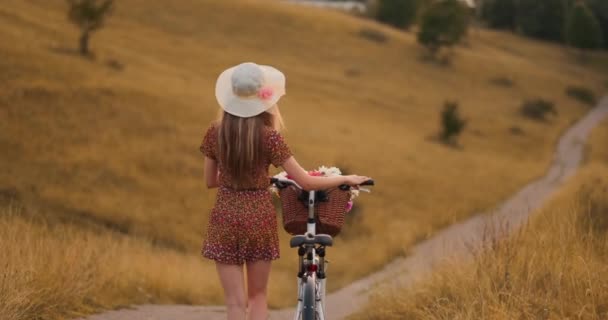 Усміхнена щаслива дівчина в сукні і капелюсі їде ретро-велосипед в парку і дивиться на камеру — стокове відео