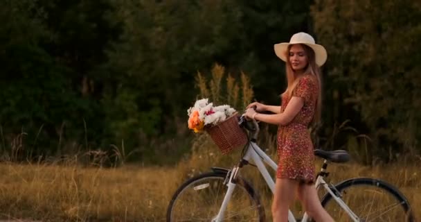 Zijaanzicht: glimlachend meisje in een jurk wandelen met een fiets in de zomer op de Stuur mand met wilde bloemen. — Stockvideo