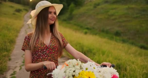 Porter meisje wandelen op de camera in een jurk met bloemen in een mand en een fiets in het veld. — Stockvideo