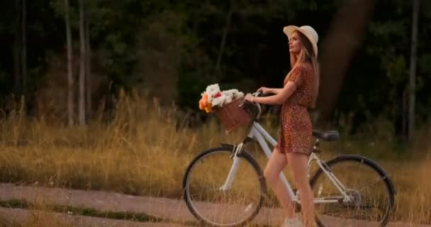 Ung leende blondin i hatt och klänning gå i klänning med cykel och blommor i korg. — Stockvideo