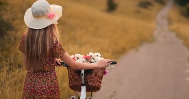 Uma menina de chapéu com uma bicicleta vira-se e olha para a câmera sorrindo . — Vídeo de Stock