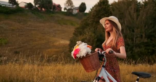 Jeune blonde souriante en chapeau et robe marchant en robe avec vélo et fleurs dans le panier . — Video