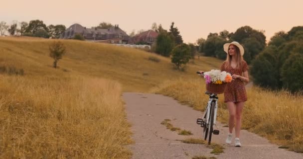 Jeune blonde souriante en chapeau et robe marchant en robe avec vélo et fleurs dans le panier . — Video