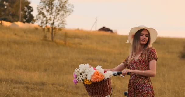 Porter dziewczyna chodzenie na aparat fotograficzny w sukni z kwiatami w koszyku i rower w polu. — Wideo stockowe