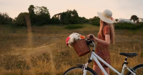 Vista lateral: niña sonriente en un vestido caminando con una bicicleta en el verano en la canasta del manillar con flores silvestres . — Vídeos de Stock