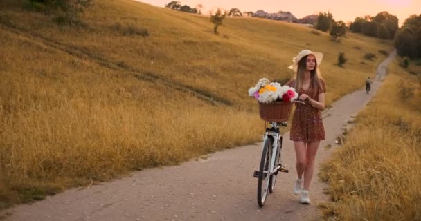 Uma mulher bonita com uma bicicleta em um chapéu e um vestido de verão leve vem com flores em uma cesta e sorri . — Vídeo de Stock