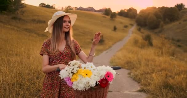 Porter meisje wandelen op de camera in een jurk met bloemen in een mand en een fiets in het veld. — Stockvideo