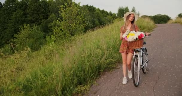 Une belle femme avec un vélo dans un chapeau et une robe d'été légère est livré avec des fleurs dans un panier et sourit . — Video