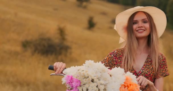 Chica de plan medio en vestido va con bicicleta y flores en el campo . — Vídeo de stock