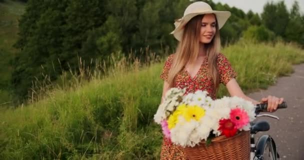 Porter meisje wandelen op de camera in een jurk met bloemen in een mand en een fiets in het veld. — Stockvideo