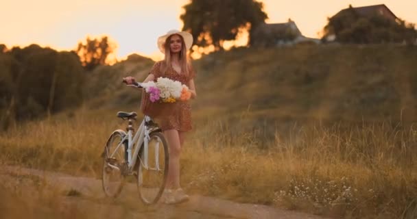 Jeune blonde souriante en chapeau et robe marchant en robe avec vélo et fleurs dans le panier . — Video