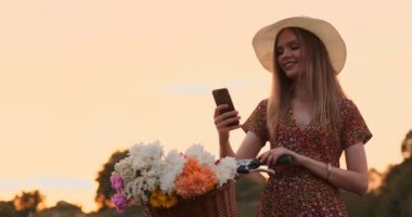 Cep telefonu ekranına ve gidonüzerinde çiçeklerle bir sepet bakarak bir bisiklet ile şapka güzel sarışın.