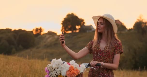 Mooie blonde in een hoed met een fiets kijken naar het scherm van de mobiele telefoon en een mand op het stuur met bloemen. — Stockvideo