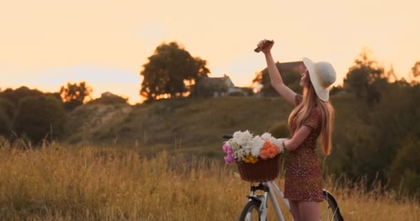 Mooie blonde in een hoed met een fiets kijken naar het scherm van de mobiele telefoon en een mand op het stuur met bloemen. — Stockvideo