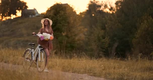 Ung vacker blond flicka i sommar i klänning och hatt går på väg med cykel och blommor i slow motion — Stockvideo