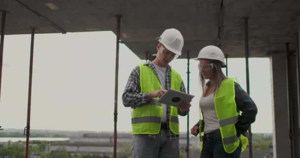 Trabalhador da construção homem e mulher arquiteto em um capacete, discutir o plano de construção da casa, dizer uns aos outros sobre o projeto, segurando um tablet, olhar para os desenhos, fundo dos raios de sol — Vídeo de Stock