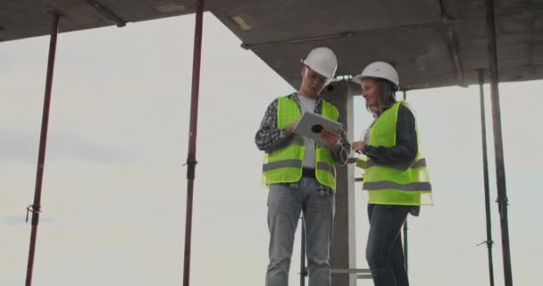 Dos ingenieros un hombre y una mujer con una tableta en un sitio de construcción condenan el plan de construcción del edificio — Vídeos de Stock