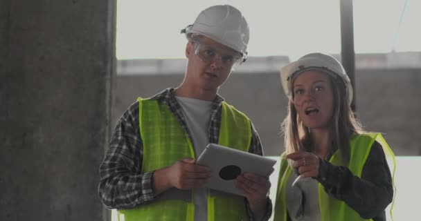 Dos ingenieros un hombre y una mujer con una tableta en un sitio de construcción condenan el plan de construcción del edificio — Vídeos de Stock