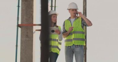 Bina planını görselleştirmek için tablet ve cep telefonu kullanan bir kadın ve erkek mühendis ile inşaat halinde bina.