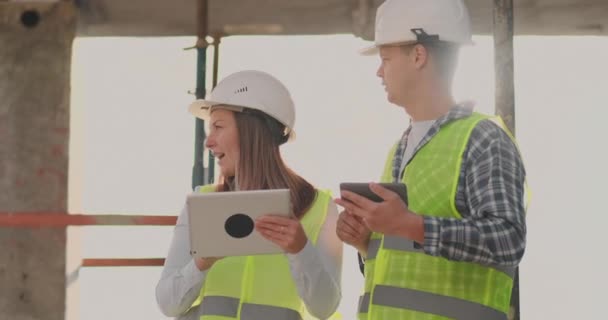 Ingenieure stehen auf dem Dach des im Bau befindlichen Gebäudes und diskutieren Plan und Baufortschritt mit Tablet und Handy — Stockvideo