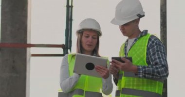 Bina planını görselleştirmek için tablet ve cep telefonu kullanan bir kadın ve erkek mühendis ile inşaat halinde bina.