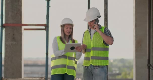 Edificio en construcción con una mujer y un hombre constructores, constructores, ingenieros caminando a lo largo de ella . — Vídeos de Stock