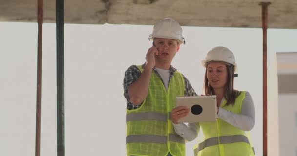Edificio en construcción con una mujer y un hombre ingenieros utilizando una tableta y teléfono móvil para visualizar el plan de construcción . — Vídeos de Stock