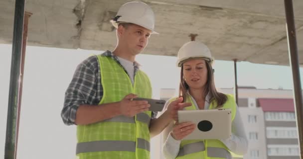 Edificio en construcción con una mujer y un hombre ingenieros utilizando una tableta y teléfono móvil para visualizar el plan de construcción . — Vídeos de Stock