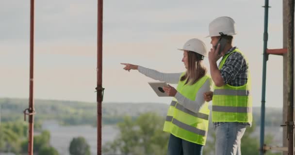 Ingenieurs ontwerpers staan op het dak van het gebouw in aanbouw en bespreken het plan en de voortgang van de bouw met behulp van een Tablet en mobiele telefoon — Stockvideo