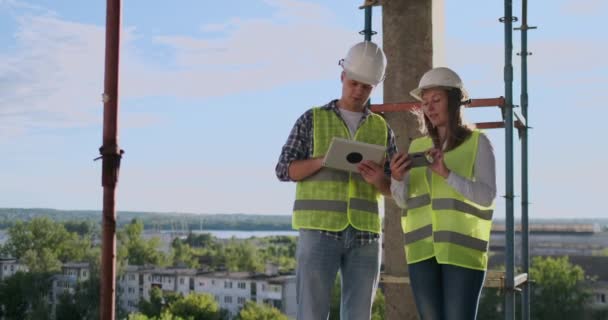 Engenheiros construtores homem e mulher de pé no telhado do edifício com um computador tablet discutindo em capacetes brancos e camisas . — Vídeo de Stock