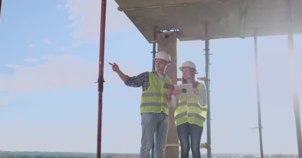 Engenheiros construtores homem e mulher de pé no telhado do edifício com um computador tablet discutindo em capacetes brancos e camisas . — Vídeo de Stock