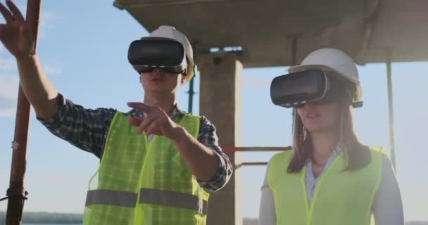 Un hombre y una mujer ingenieros en un sitio de construcción en gafas VR manejan la construcción de un edificio discutiendo un plan de desarrollo . — Vídeos de Stock