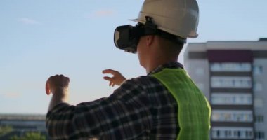 Gün batımında binanın çatısında mühendis Builder VR gözlük standları ve geleceğin arayüzünü kullanarak ellerini taşır. Geleceğin fütüristik mühendisi. Arka görünüm.