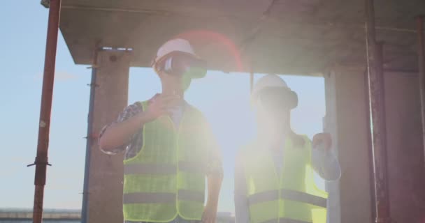 Un hombre y una mujer ingenieros en un sitio de construcción en gafas VR manejan la construcción de un edificio discutiendo un plan de desarrollo . — Vídeos de Stock