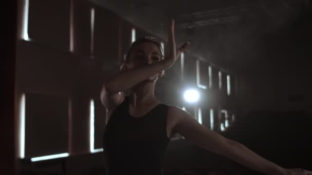 Изящная балерина в темном платье на темной сцене театра в дыму исполняет танцевальные движения в замедленной съемке — стоковое видео