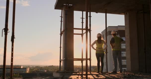 Dos Ingeniero Jefe con un dibujo en la mano mirando el sitio de construcción. Vista desde atrás. Ingeniero jefe con el plano en la mano mirando la construcción . — Vídeos de Stock