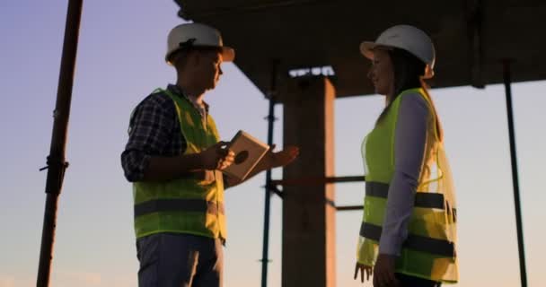Construtores homem com um tablet e uma mulher de capacetes brancos apertar as mãos ao pôr do sol de pé no telhado do edifício . — Vídeo de Stock