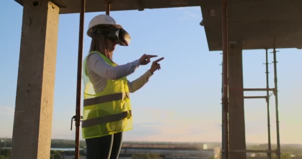 Ispettore donna in VR occhiali e casco controlla i progressi della costruzione di un grattacielo muovendo le mani al tramonto visualizzando il piano dell'edificio . — Video Stock