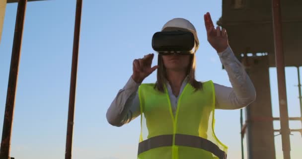 Mujer inspector en gafas VR y casco comprueba el progreso de la construcción de un rascacielos moviendo las manos al atardecer visualizando el plan del edificio . — Vídeo de stock