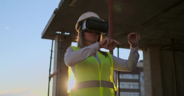 Mujer experta en ingeniería Constructora en gafas VR y casco comprueba el progreso de la construcción de rascacielos moviendo sus manos al atardecer visualizando el plan de construcción . — Vídeo de stock