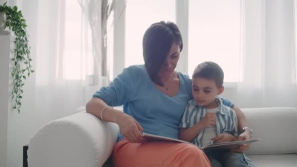 Мать и сын читают книги. Счастливая молодая мать и сын читают книги на диване дома . — стоковое видео