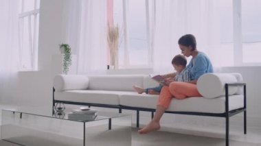 Kanepede oturma odasında evin parlak beyaz iç oturan bir kitap okuma bir çocuk ile genç bir anne.