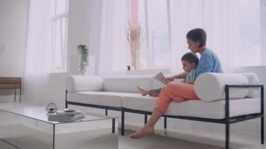 Kanepede oturma odasında evin parlak beyaz iç oturan bir kitap okuma bir çocuk ile genç bir anne.