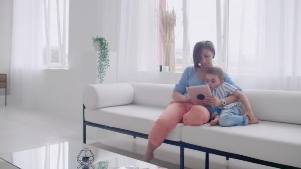 Mor och son sitter på soffan med digital tablett. Glad mamma och liten pojke med hjälp av surfplatta med pekskärm tillsammans titta på en video. Leende mor och söt pojke spelar på digitala tablett — Stockvideo