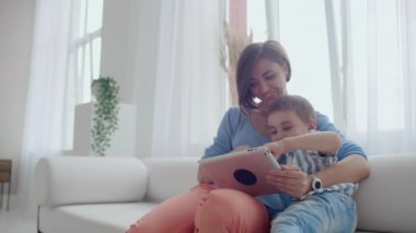 Anne ve oğlu dijital tablet kullanarak koltukta oturuyor. Tablet dokunmatik ekranlı bir video izlerken birlikte mutlu anne ve küçük çocuk kullanarak. Gülümseyen anne ve sevimli çocuk dijital tablet üzerinde oynama