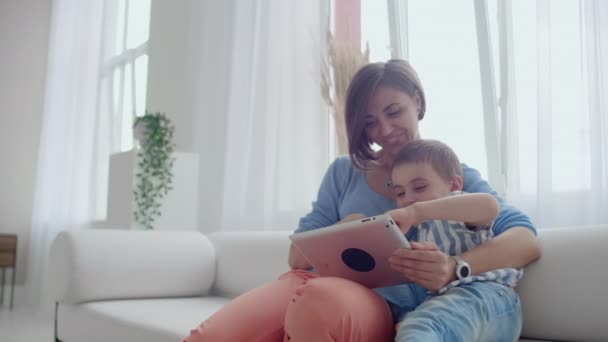 Madre e hijo sentados en un sofá usando una tableta digital. Feliz mamá y el niño usando la tableta con pantalla táctil juntos viendo un video. Sonriente madre y lindo niño jugando en la tableta digital — Vídeos de Stock