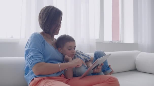 Ευτυχισμένος οικογένεια μαμά και παιδί γιός χρησιμοποιώντας ψηφιακό δισκίο κάθεται στον καναπέ, χαμογελαστή μητρική μητέρα με το παιδί γιος κρατώντας PC υπολογιστή κοιτάζοντας στην οθόνη κάνουν online αγορές κάνουν βίντεο κλήση ρολόι κινούμενα σχέδια. — Αρχείο Βίντεο